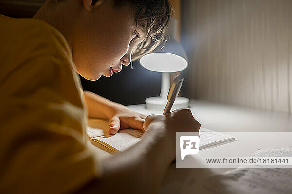 Junge schreibt zu Hause unter beleuchteter Lampe in ein Buch