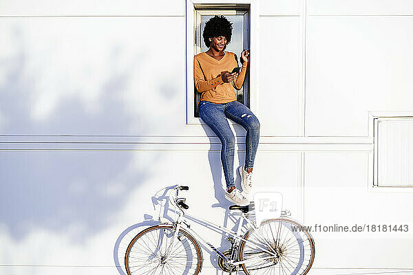 Lächelnde Frau mit Fahrrad und Mobiltelefon auf der Fensterbank