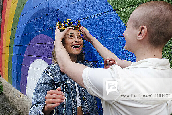 Non-binary person adjusting crown on lesbian woman's head by rainbow wall