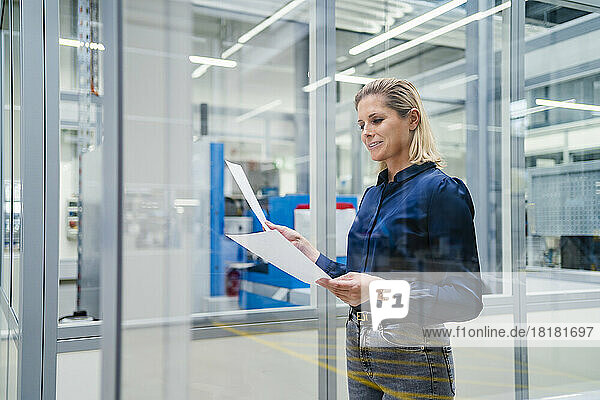 Smiling businesswoman reviewing documents in factory seen through glass