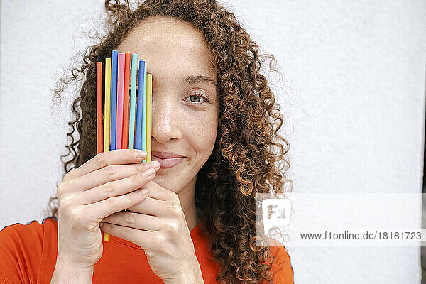 Smiling woman covering eye with colored pencils in front of white wall