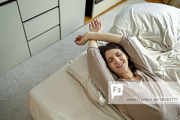 Lächelnde Frau mit erhobenen Händen  die zu Hause im Bett aufwacht