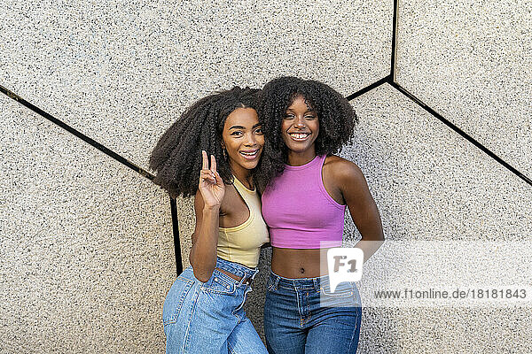 Smiling woman gesturing peace sign standing with friend in front of wall