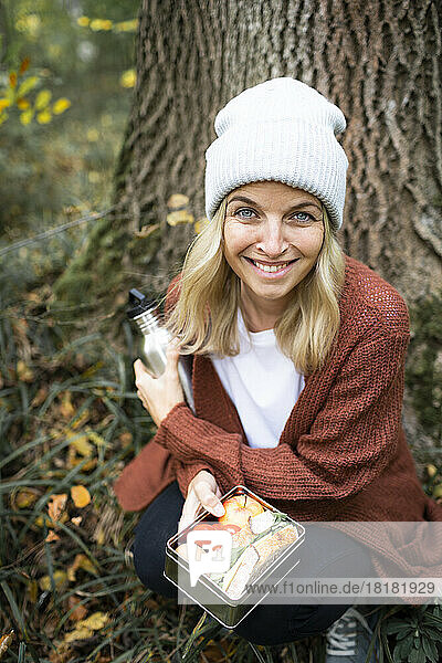 Lächelnde Frau mit Lunchbox hockt am Wochenende im Wald