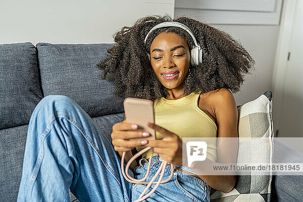 Smiling woman using smart phone and listening to music through wireless headphones at home