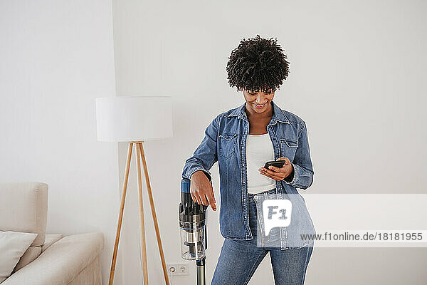 Smiling woman standing with vacuum cleaner and using smart phone at home
