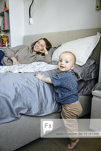 Cute boy standing by bed with mother in background