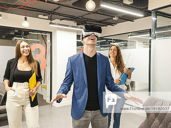 Glücklicher Geschäftsmann mit VR-Brille mit Kollegen im Hintergrund im Büro