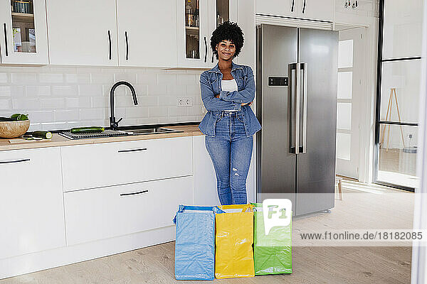 Smiling woman with arms crossed standing by reusable bags in kitchen at home