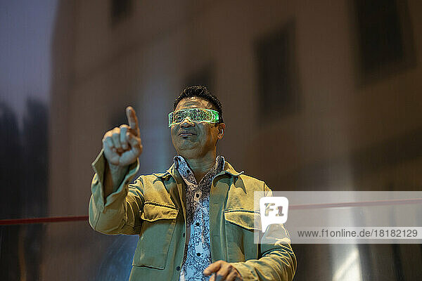 Smiling man wearing smart glasses gesturing in front of building at night