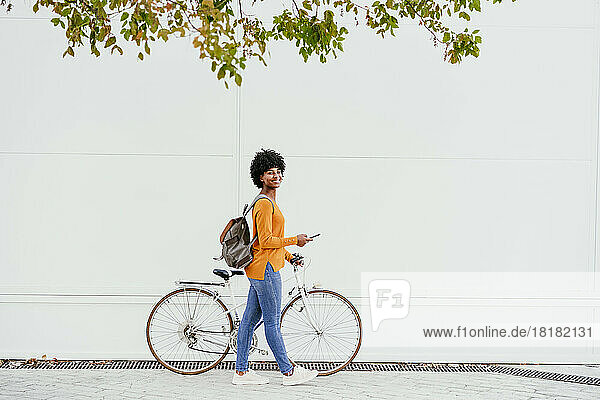 Smiling woman with backpack and smart phone wheeling bicycle by wall