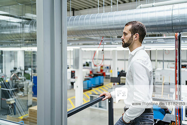 Serious young businessman with hand in pocket standing by railing in factory