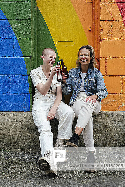 Happy young woman toasting beer with non binary friend sitting in front of colorful wall