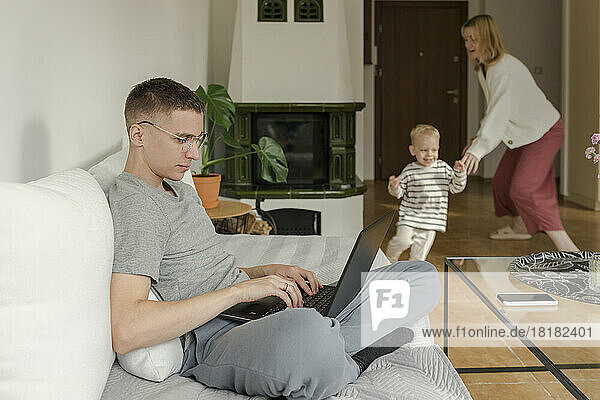 Young man using laptop with mother and son playing in background