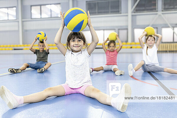 Smiling girl holding ball overhead sitting with friends at school sports court