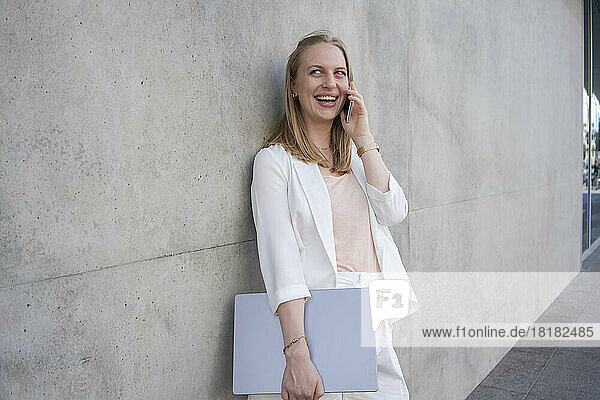 Smiling freelancer with laptop talking on smart phone by concrete wall