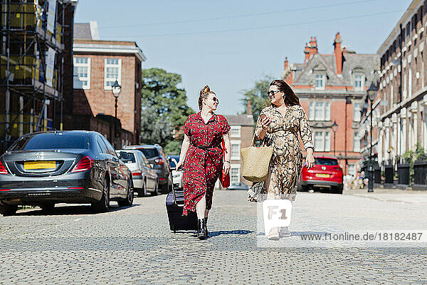 Zwei Freunde  die mit Rollgepäck und Einkaufstasche auf der Straße spazieren und einander anschauen  Liverpool  Großbritannien