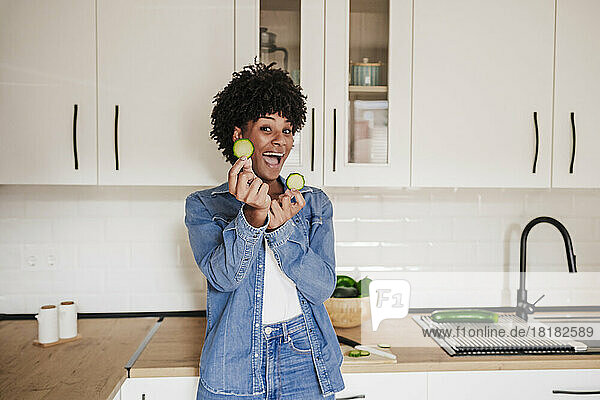 Fröhliche junge Frau zeigt Zucchinischeiben und lehnt zu Hause an der Küchentheke