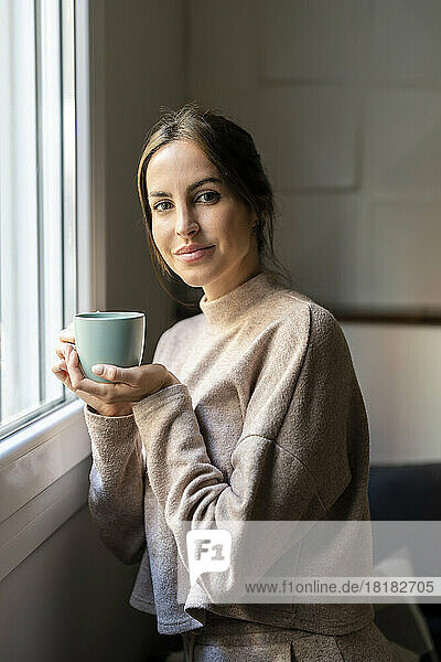 Smiling woman with cup of tea standing by window at home