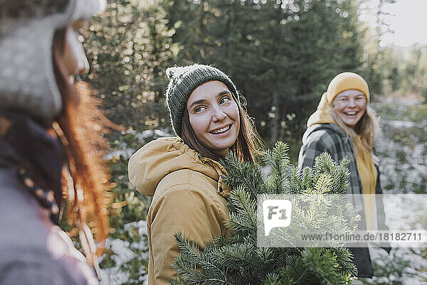Smiling woman with spruce tree twigs looking at friend in forest