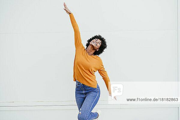 Happy woman dancing with arms raised in front of wall