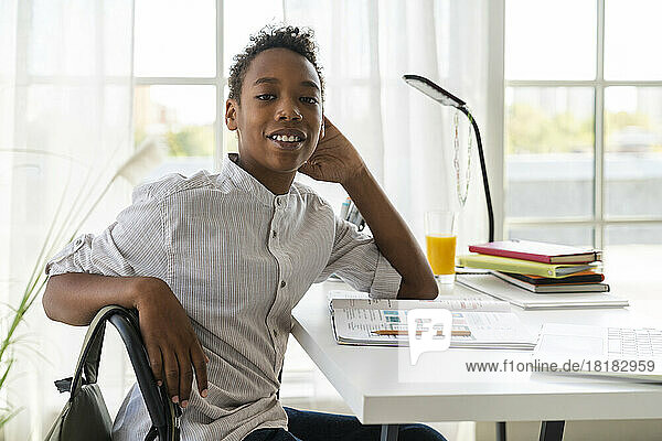 Lächelnder Junge sitzt zu Hause auf einem Stuhl mit Lehrbuch