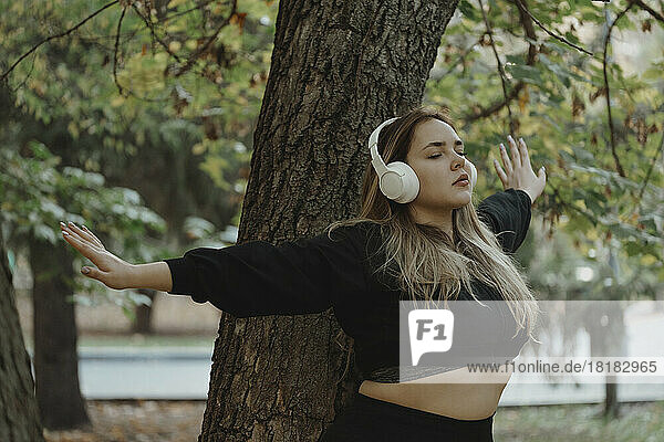 Young woman listening to music through wireless headphones standing with arms outstretched leaning on tree trunk