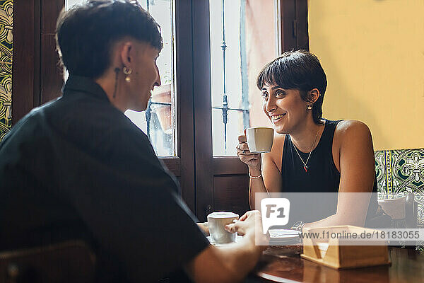 Glückliche junge Frau hält Kaffeetasse in der Hand und unterhält sich mit ihrem Freund im Café