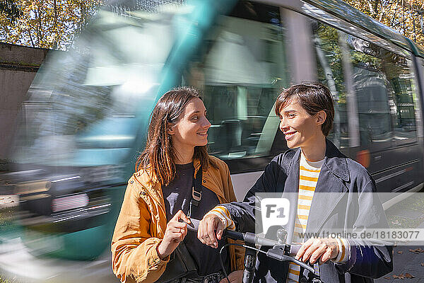 Smiling woman looking at friend standing with push scooter by passing tram