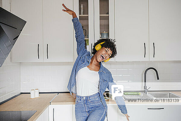 Cheerful young woman wearing wireless headphones dancing in kitchen at home