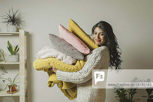 Smiling young woman holding pillows and blankets by wall at home