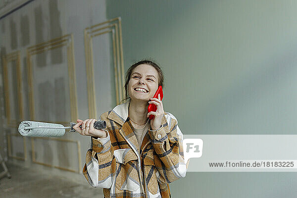 Happy woman with paint roller talking on smart phone in apartment