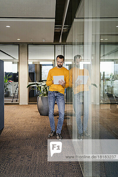 Young businessman using tablet PC leaning on glass wall in office