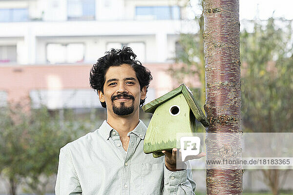 Smiling man holding green birdhouse by tree trunk