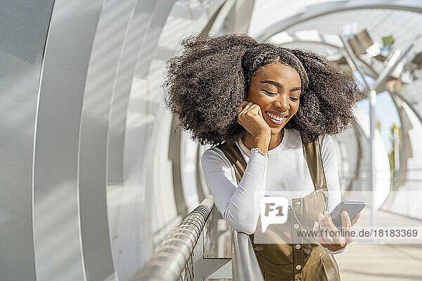 Smiling woman using smart phone leaning on bridge railing
