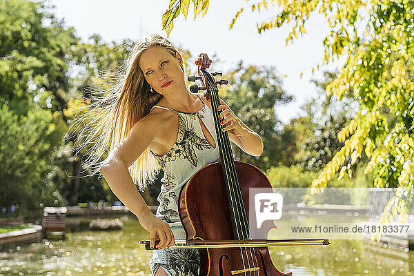 Blond woman playing cello in front of lake at park