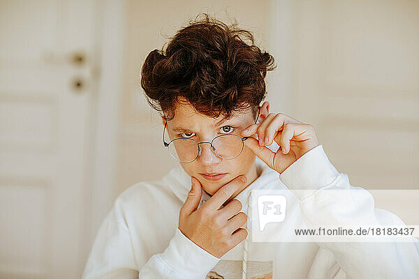 Boy adjusting eyeglasses with hand on chin at home