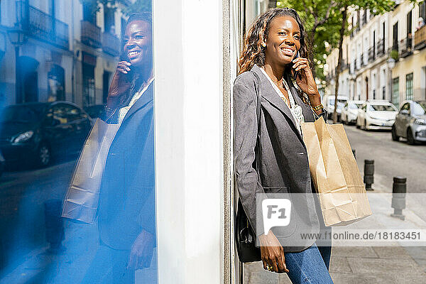 Smiling woman carrying shopping bags talking through mobile phone leaning on wall