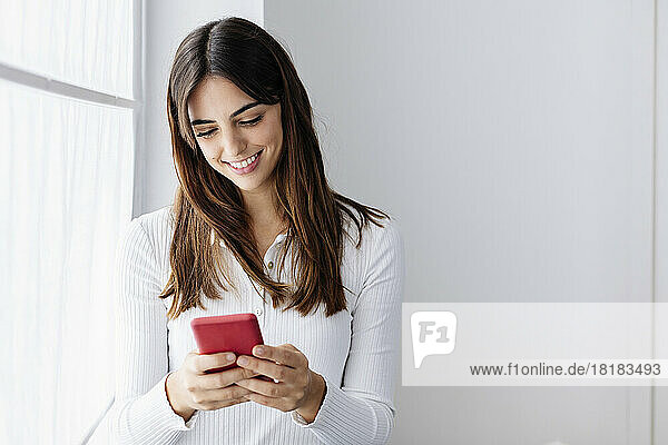 Smiling woman using smart phone by window at home