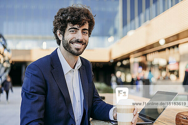 Smiling businessman with laptop and disposable coffee cup sitting in cafe