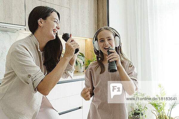 Happy girl with mother singing into pepper mills and enjoying in kitchen