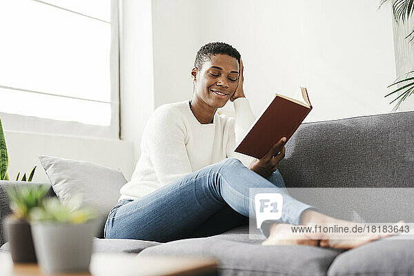 Smiling woman with closed eyes sitting on couch at home holding book