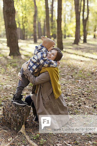 Boy standing on tree stump embracing mother in park