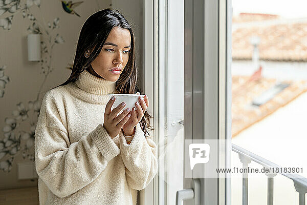 Thoughtful young woman standing with bowl of soup by window at home