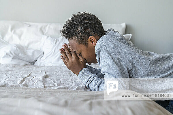 Boy with hands clasped leaning on bed at home
