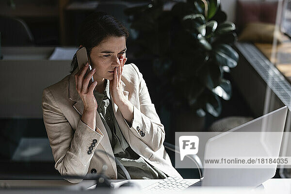 Gestresste Geschäftsfrau  die am Schreibtisch im Büro mit dem Handy telefoniert und durch Glas gesehen wird