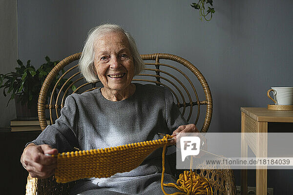 Smiling senior woman with knitting wool and needle on chair at home