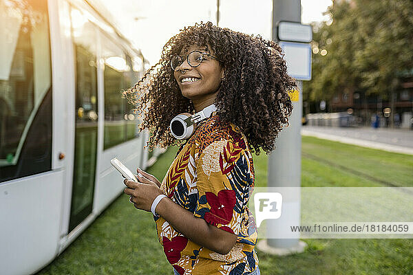 Happy young woman standing with smart phone by tram