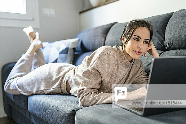 Junge Frau schaut sich einen Film auf einem Laptop an  der zu Hause auf der Couch liegt