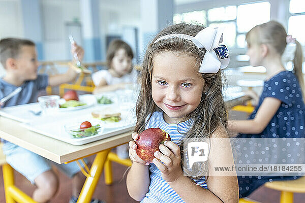 Smiling girl holding apple sitting with friends in school cafeteria